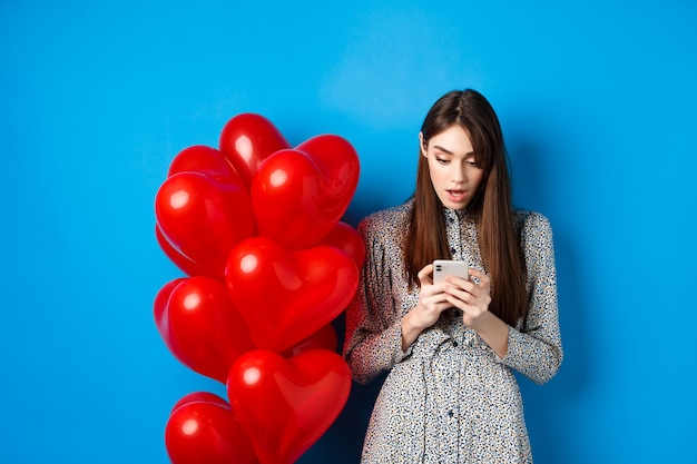 San Valentino. Ritratto di giovane donna in piedi vicino a palloncini romantici rossi, guardando sorpreso lo schermo dello smartphone, sfondo blu