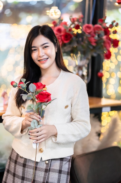 San Valentino Ritratto felice sorridente bella giovane donna asiatica che indossa abiti caldi con in mano un mazzo di rose rosse con vaso di rose nel soggiorno all'interno o caffè