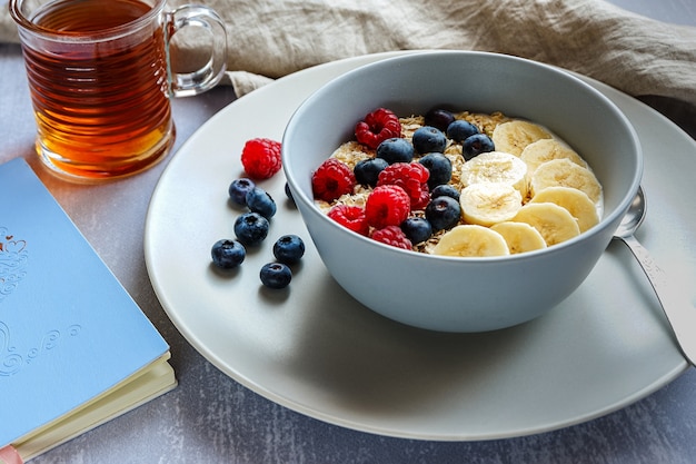 Sana colazione con farina d'avena in una ciotola, fette di banana, lampone, mirtillo, una tazza di tè e un taccuino sul piano del tavolo grigio chiaro