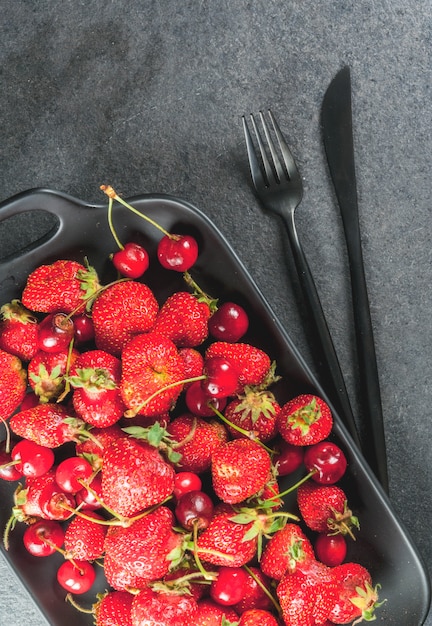 Sana colazione, pranzo o merenda. Frutti e bacche estive. Piatto rettangolare organico di ciliegie e fragole fresche, con una forchetta, su un tavolo di pietra nera.