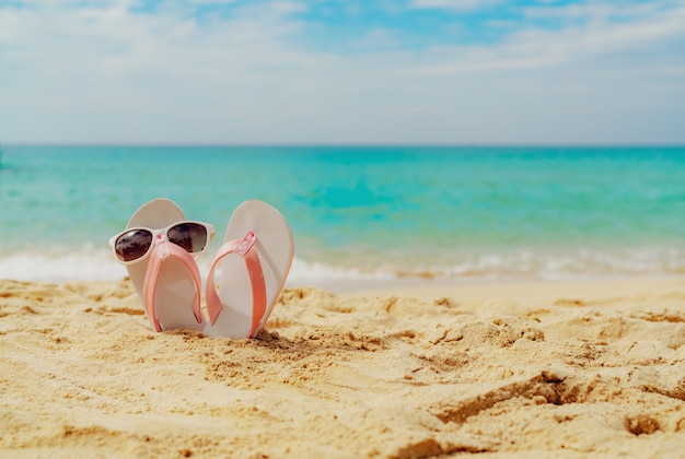 Sandali rosa e bianchi, occhiali da sole sulla spiaggia di sabbia in riva al mare. Infradito stile casual e occhiali in riva al mare. Vacanze estive sulla spiaggia tropicale. Divertente viaggio di vacanza sulla spiaggia di sabbia.