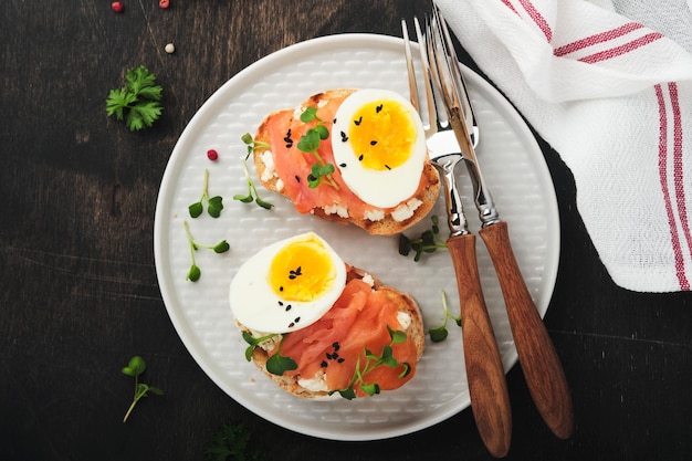 Sandwich con condimenti deliziosi salmone affumicato uova erbe e microgreens ravanello nero semi di sesamo su piatto bianco su vecchio tavolo in legno sfondo Sano panino aperto superfood Vista dall'alto