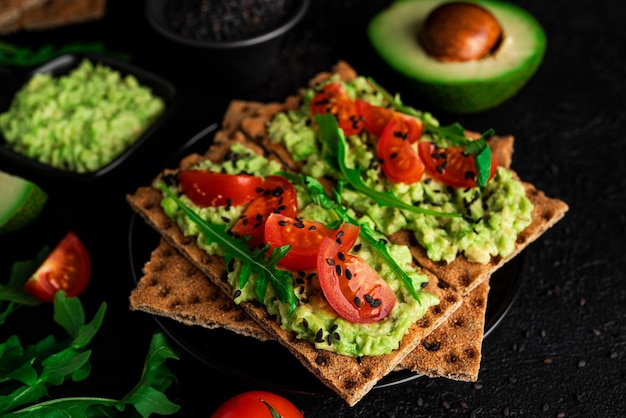 Sandwich di avocado con crema di avocado e pane croccante di segale per uno spuntino. Fibre, fitness e cibo dietetico. Pane di segale con guacamole, rucola e pomodorini su fondo scuro. Foto di alta qualità