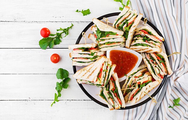 Sandwich di club con prosciutto, pomodoro, formaggio al cetriolo e arugula su sfondo di legno