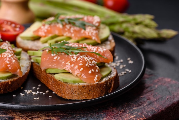 Sandwich di pane tostato con burro di avocado e salmone decorato con rucola e semi di sesamo su fondo di pietra nera