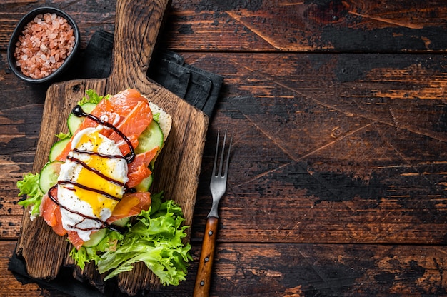 Sandwich di salmone affumicato con uovo alla Benedict e avocado su pane. Fondo in legno scuro. Vista dall'alto. Copia spazio.