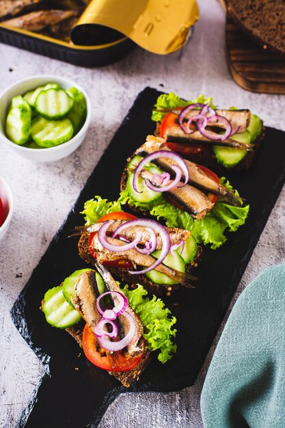 Sandwich su pane di segale con sprat affumicati verdure e erbe vegetali su ardesia vista verticale