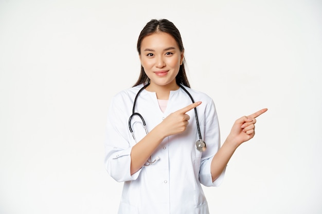 Sanità, concetto di persone mediche. Sorridente dottoressa asiatica, puntando le dita a destra, mostrando pubblicità, informazioni sull'ospedale, in piedi su sfondo bianco