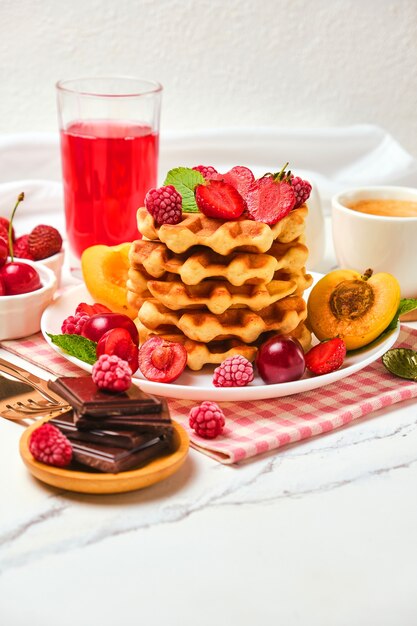 Sano set colazione con cialde belghe con fragole, albicocche, ciliegie, succo di frutta e una tazza di caffè nero e cioccolato amaro su sfondo tavolo in pietra bianca