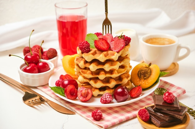 Sano set colazione con cialde belghe con fragole, albicocche, ciliegie, succo di frutta e una tazza di caffè nero e cioccolato amaro su sfondo tavolo in pietra bianca