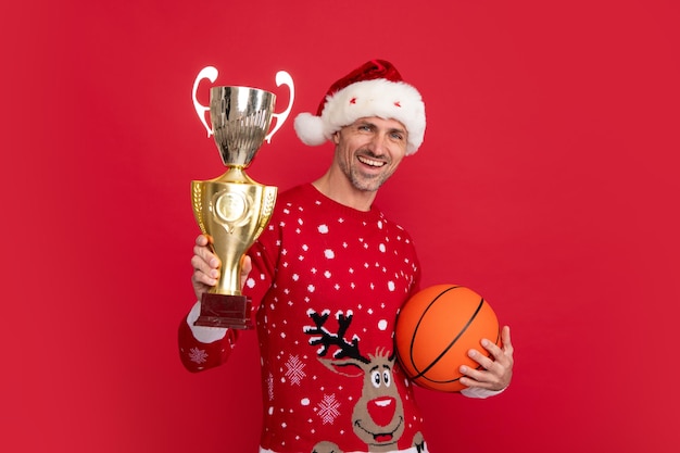 Santa trainer tenere palla da basket e coppa campione Ritratto di uomo di mezza età in maglione isolato su sfondo rosso Concetto di vacanze felicità emozioni e celebrazione del Natale