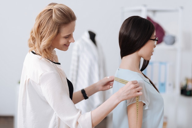 Sarto femminile in piedi presso l'atelier, tenendo un nastro e misurando la schiena di una donna bruna con gli occhiali.