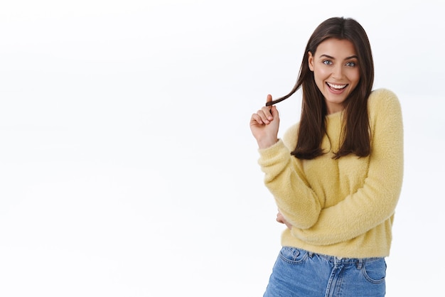 Sassy bella giovane donna moderna elegante in maglione giallo che è civettuola incontrare qualcuno e flirtare sorridente ciocca di capelli rotolante sfacciata a portata di mano e guardando la fotocamera audace sfondo bianco