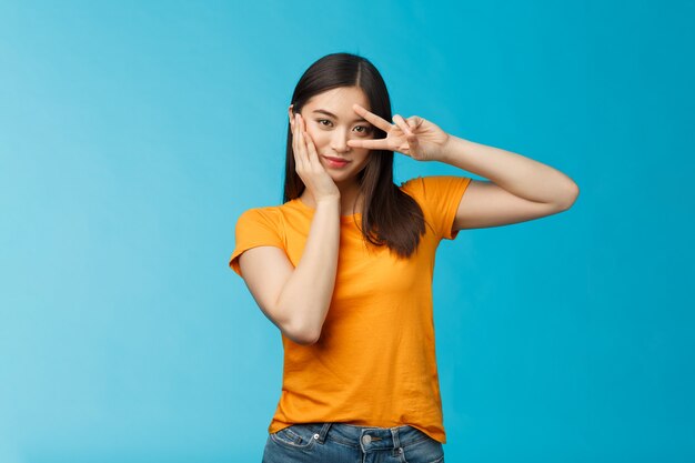 Sassy elegante bella ragazza asiatica assertiva coreana mostra il segno della vittoria della pace sull'occhio, tocca la guancia sembra autorizzata e motivata, in piedi sfacciato sfondo blu civettuolo, indossa una t-shirt gialla