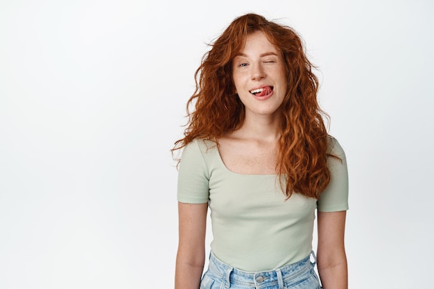 Sassy giovane donna rossa con capelli lunghi naturali che strizza l'occhio e mostra la lingua che sorride alla telecamera in piedi in maglietta su sfondo bianco
