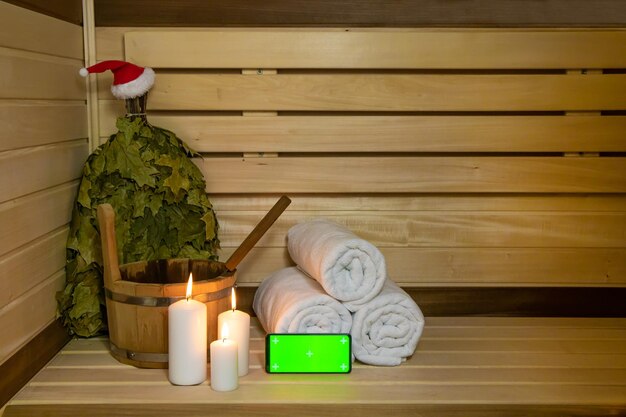 Sauna. Telefono con schermo verde, candele, asciugamano, secchio di legno, cappello da Babbo Natale e scopa, copia spazio, testo,