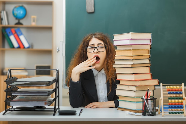 sbadigliando giovane insegnante femminile che indossa occhiali seduto alla scrivania con strumenti scolastici in classe