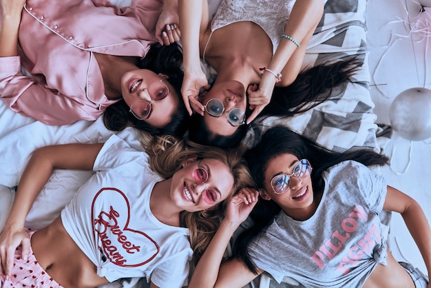 Sbrigati ad essere felice. Vista dall'alto di quattro giovani donne giocose in pigiama e occhiali che sorridono mentre sono sdraiate sul letto a casa