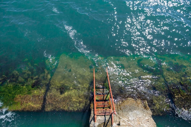 Scala di metallo rossa per la discesa in mare ricoperta di alghe contro l'acqua blu