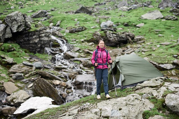 Scalatore castana femminile che cammina con i bastoni da passeggio in mani su un pendio roccioso verde davanti alle belle montagne rocciose