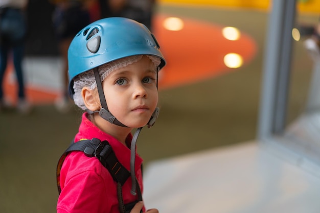 Scalatore della bambina sveglia in casco protettivo blu e attrezzatura per l'arrampicata in piedi nel parco divertimenti del centro scalatore per bambini