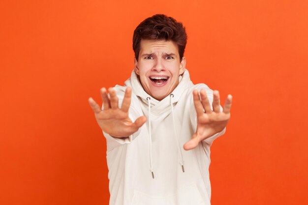 Scaorange uomo in maglione leggero casual urlando tenendo le mani davanti alla telecamera panico sul viso adolescente paura delle allucinazioni di fantasmi Colpo in studio al coperto isolato su sfondo arancione