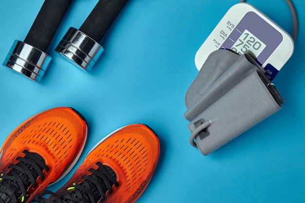 Scarpe da ginnastica arancioni, manubri e monitor della pressione sanguigna su sfondo blu. Esercizio e pressione sanguigna