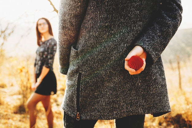 Scatola anello nascosto maschile irriconoscibile dietro la schiena mentre si trova vicino a una donna sfocata in giornata di sole in campagna