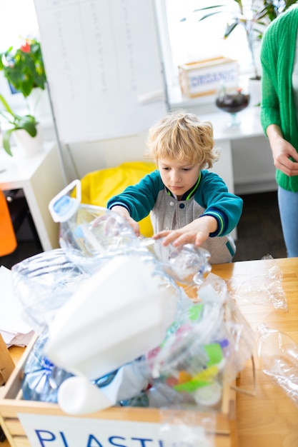 Scatola con plastica. Scolaro bello dai capelli biondi in piedi vicino a una scatola con plastica dopo aver ordinato i rifiuti