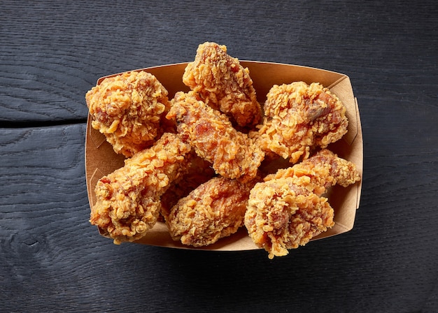 Scatola di carta da asporto di ali di pollo fritte su fondo di legno nero, vista dall'alto