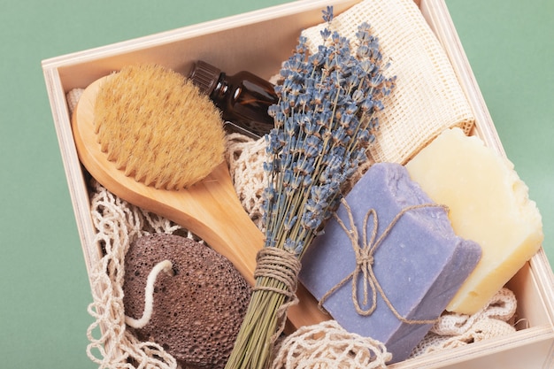 Scatola di legno con accessori cosmetici riutilizzabili su sfondo verde. Sapone organico, spazzola da massaggio in legno, pietra pomice, bottiglia di vetro