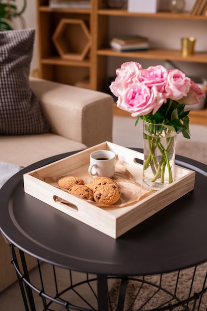 Scatola di legno quadrata con mazzo di rose rosa fresche in vetro, tazza di caffè e biscotti sul piccolo tavolo rotondo in ambiente domestico