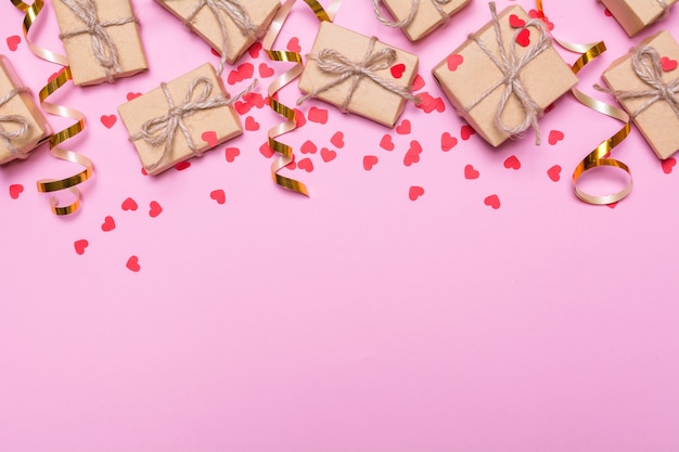 Scatole regalo avvolte in carta kraft su sfondo rosa. Cuori di coriandoli e nastri d'oro, decorazioni festive. Design piatto laici, vista dall'alto