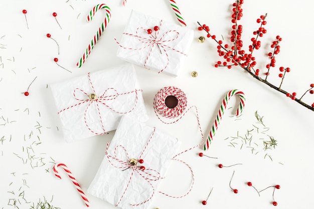 Scatole regalo di Natale con caramelle festive, ramo con bacche rosse, bobina di corda di Natale e aghi di abete sul tavolo bianco. Vista piana laico e dall'alto