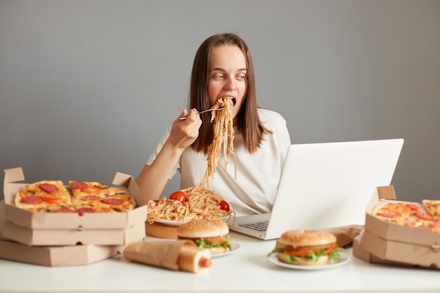 Scatto al coperto di donna affamata indaffarata che mangia fast food mentre lavora al laptop seduto al tavolo contro il muro grigio lavorando al laptop e mangiando pasta tiene la bocca aperta guardando il display del notebook