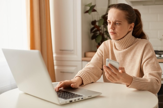 Scatto al coperto di una donna caucasica concentrata che lavora sodo che indossa un maglione beige in stile casual con acconciatura a coda di cavallo seduta al tavolo davanti al computer portatile e tenendo in mano lo smartphone.