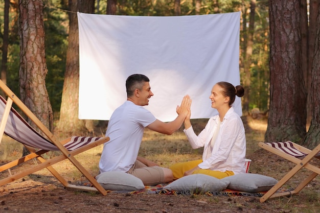 Scatto all'aperto di una giovane coppia positiva seduta nella foresta con una lavagna luminosa con schermo bianco vuoto con la famiglia dell'area pubblicitaria che si tiene per mano o dà il cinque