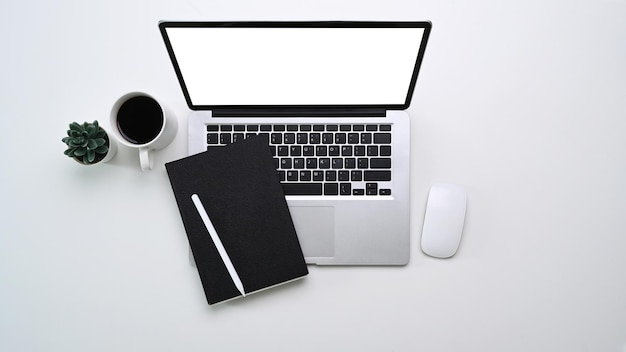 Scatto dall'alto del moderno ambiente di lavoro minimale con computer portatile, notebook, tazza di caffè e pianta su sfondo bianco.