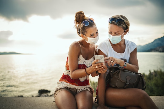 Scatto di amiche felici con maschera protettiva che trascorrono del tempo in riva al mare e navigano sui social media durante l'esplorazione di un Mediterraneo durante la pandemia della corona.