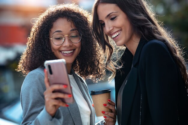 Scatto di due belle donne d'affari che usano lo smartphone mentre bevono caffè passeggiando per la città.