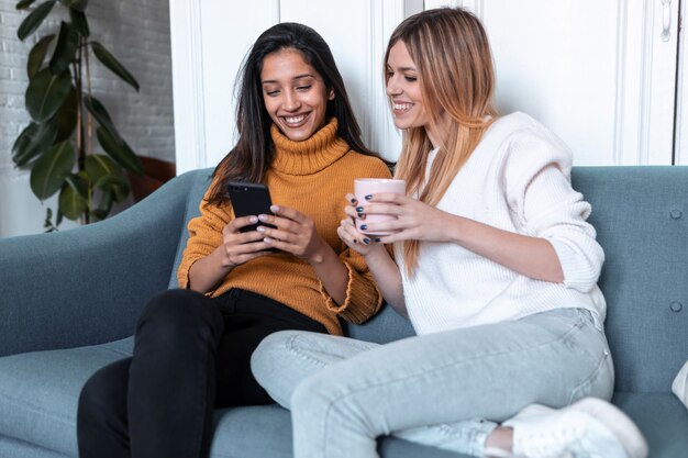 Scatto di due belle giovani donne che usano il cellulare mentre bevono caffè sul divano di casa.