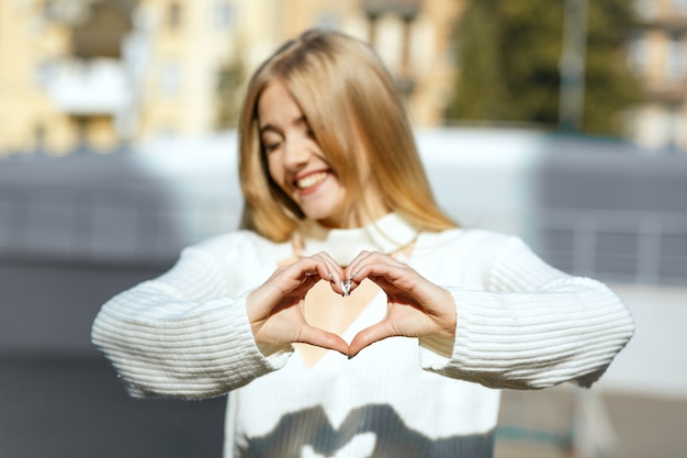 Scatto di stile di vita: ragazza bionda espressiva in maglione lavorato a maglia bianco che fa un simbolo del cuore con le sue mani