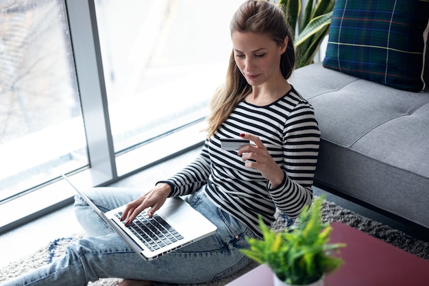 Scatto di una giovane donna che fa shopping online con carta di credito e laptop mentre è seduta per terra a casa.