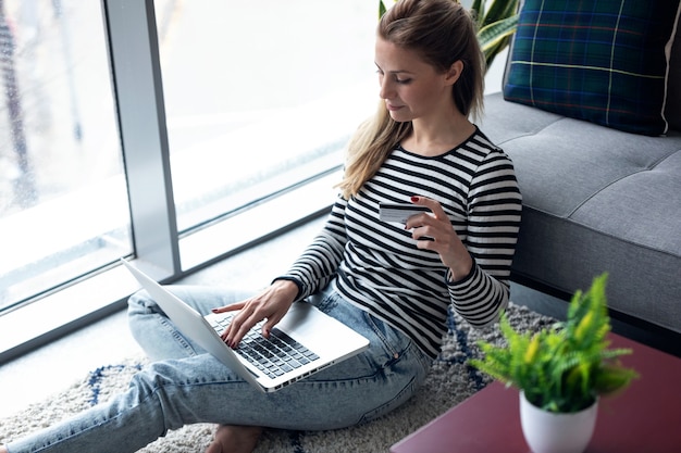 Scatto di una giovane donna che fa shopping online con carta di credito e laptop mentre è seduta per terra a casa.