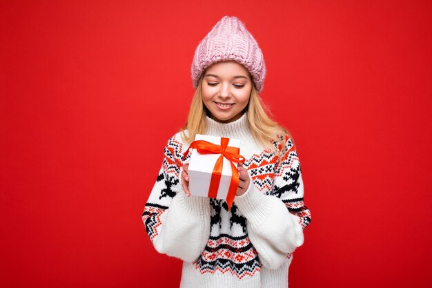 Scatto fotografico di bella giovane donna bionda sorridente felice isolata su una parete di fondo rossa che indossa un maglione invernale e un cappello rosa che tiene in mano una confezione regalo bianca con un nastro rosso e guarda la sorpresa