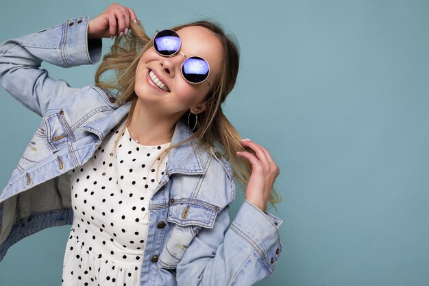 Scatto fotografico di una giovane donna bionda sorridente felice attraente che indossa abiti casual estivi e occhiali da sole eleganti isolati su una parete di sfondo colorata che guarda al lato e si diverte. copia spazio