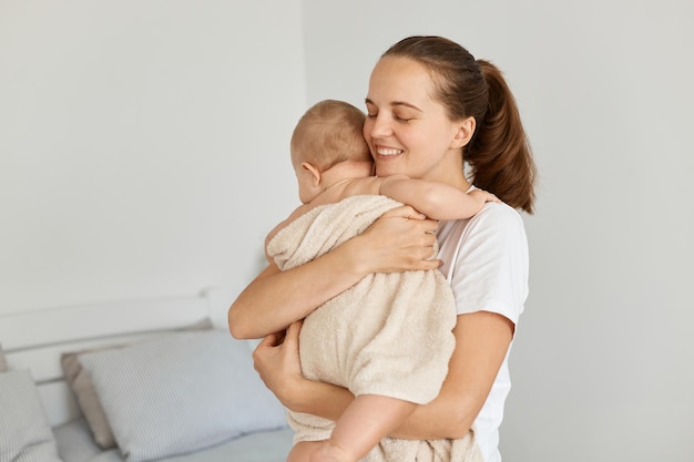 Scatto in interni di donna felice e soddisfatta che tiene la figlia avvolta in un asciugamano dopo il bagno bella madre che abbraccia il suo bambino con amore che si prende cura del bambino