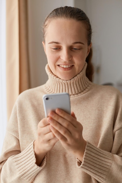 Scatto in interni di donna sorridente positiva con acconciatura a coda di cavallo che indossa un maglione beige in stile casual, in piedi con il telefono cellulare in mano, controllando i social network, scrivendo un messaggio.