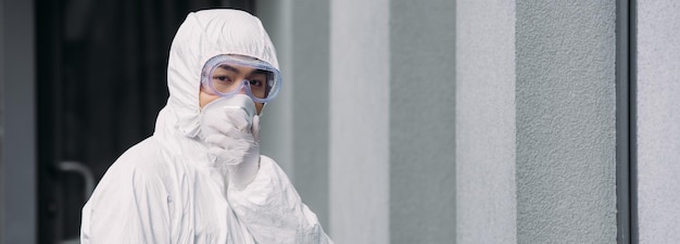 Scatto panoramico di epidemiologo asiatico in tuta ignifuga e maschera respiratoria che guarda la fotocamera mentre