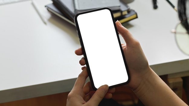 Scatto ritagliato delle mani di una donna che utilizza il modello di smartphone sullo schermo vuoto del telefono bianco della scrivania dell'ufficio